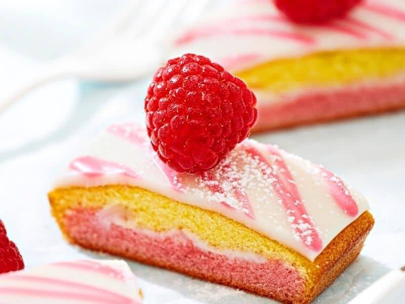 Colorful raspberry cake slices with pink and yellow layers, topped with fresh raspberries and drizzled with icing, ideal for dessert enthusiasts.