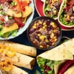 A colorful spread of Mexican food featuring tacos topped with fresh vegetables, a bowl of hearty bean and corn chili, and flour tortillas filled with greens, all arranged on a dark background.