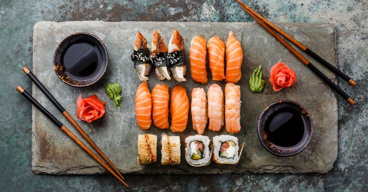 Assorted sushi platter featuring various types of nigiri, sashimi, and rolls, garnished with wasabi and pickled ginger, served on a rustic wooden board with soy sauce in small bowls and chopsticks.