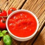 Bowl of fresh tomato sauce surrounded by ripe cherry tomatoes and basil leaves on a rustic wooden surface.