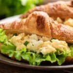 Croissant sandwich filled with scrambled eggs and fresh lettuce, served on a plate with a blurred green background.