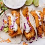 Crispy fish tacos topped with creamy sauce and purple cabbage, served with lime wedges and a side of salsa.