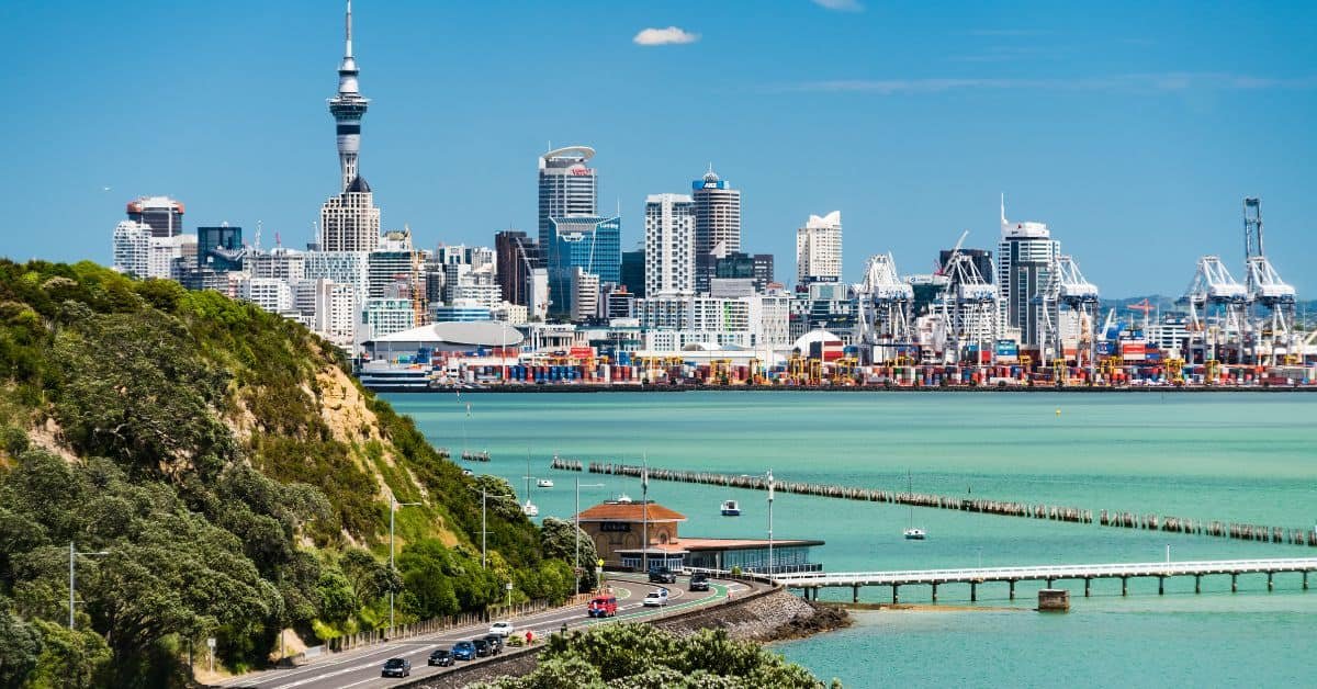 A panoramic view of Auckland's skyline featuring the Sky Tower, with a vibrant blue harbor and lush green hills in the foreground.