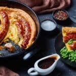 What to Serve With Toad in the Hole? Delicious toad in the hole featuring sausages in a fluffy Yorkshire pudding, served with green peas and rich gravy, set against a dark background with spices.