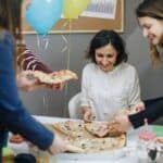 Group of friends celebrating a birthday with pizza and balloons, enjoying food together at a festive gathering.