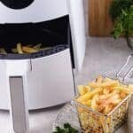 Air fryer with golden French fries in a basket, placed on a kitchen countertop with fresh herbs in the background.