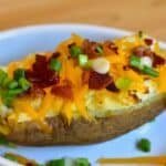 Baked potato topped with melted cheddar cheese, crispy bacon bits, and chopped green onions on a white plate.