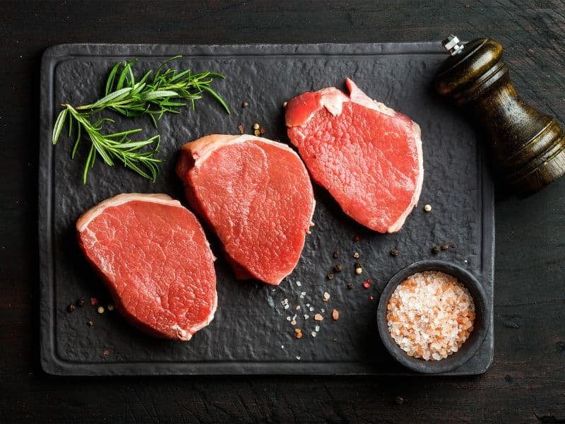 Three raw beef steaks on a dark slate surface, garnished with fresh rosemary and accompanied by a small bowl of pink salt and a pepper grinder.