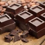 Dark chocolate squares are arranged on a wooden surface, surrounded by cocoa beans and broken chocolate pieces, highlighting the rich texture and color of premium chocolate.