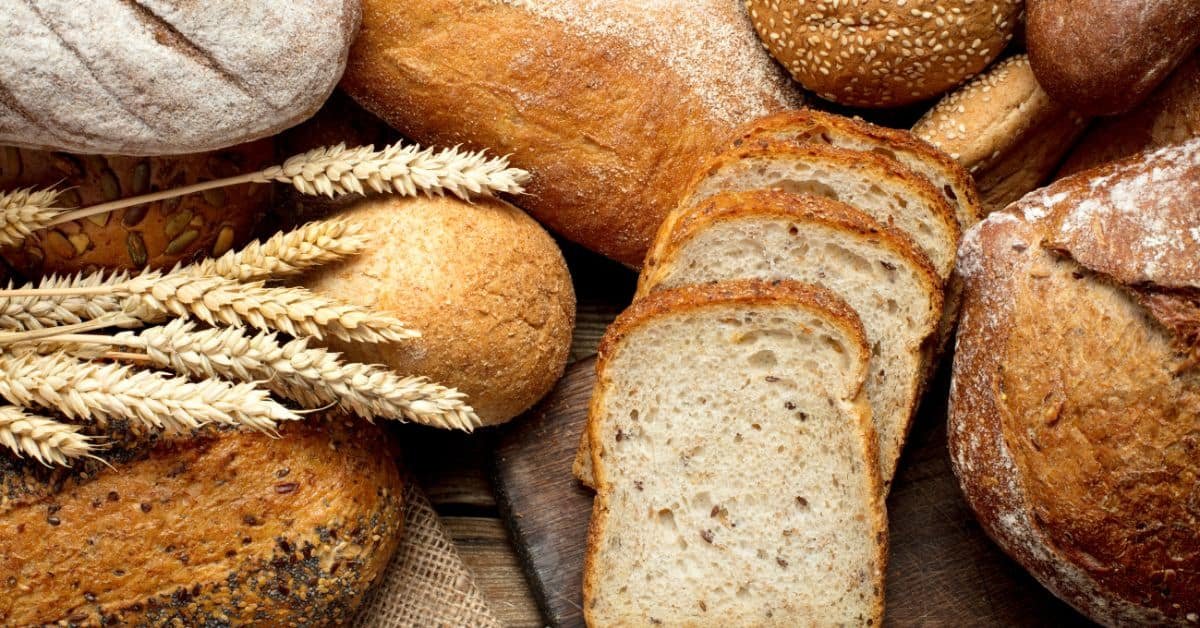 Assorted types of freshly baked bread, including whole grain, sliced loaves, and rolls, arranged with wheat stalks on a rustic wooden surface.