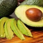 Fresh avocados on a wooden cutting board, featuring one halved avocado with a brown seed and several green slices laid beside it, showcasing their creamy texture and vibrant color.