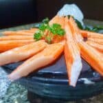 Fresh sushi platter featuring vibrant orange crab sticks arranged elegantly on a black plate, garnished with a sprig of parsley and a decorative piece of ice.