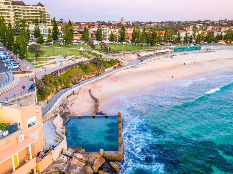 Aerial view of a beautiful beach with clear turquoise waters, sandy shores, and a pool by the ocean, surrounded by lush greenery and coastal buildings. Ideal for beachgoers and nature lovers.