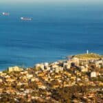 Aerial view of a coastal city with residential buildings, a green hilltop, and ships on the ocean, showcasing a serene landscape and urban development.