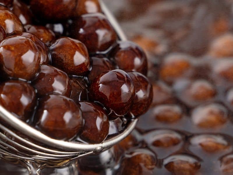 Close-up of glossy brown chocolate balls in a metal strainer, highlighting their smooth texture and rich appearance, set against a blurred background of similar chocolate treats.