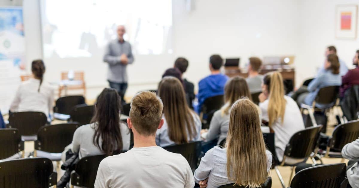 Audience listening to a speaker during a presentation in a modern classroom setting, with a focus on the back of the attendees' heads and the speaker in the foreground.