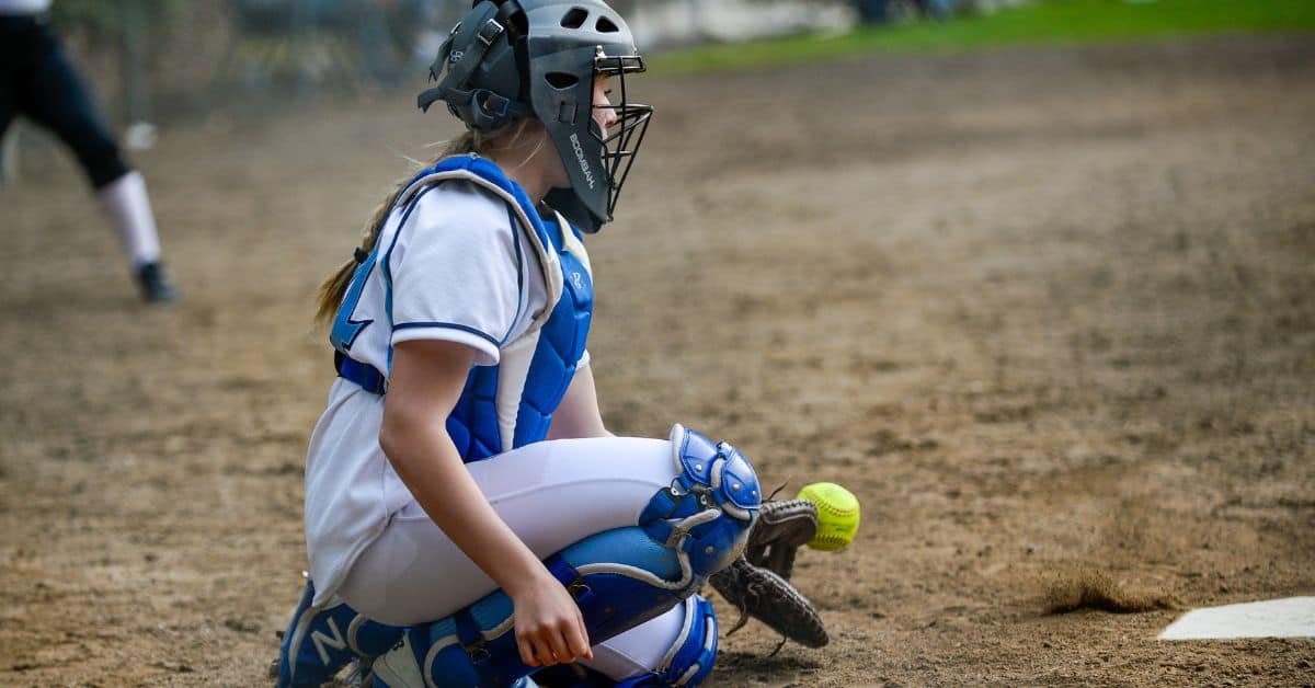 Young female softball catcher in protective gear, crouching on the field as a yellow softball approaches.