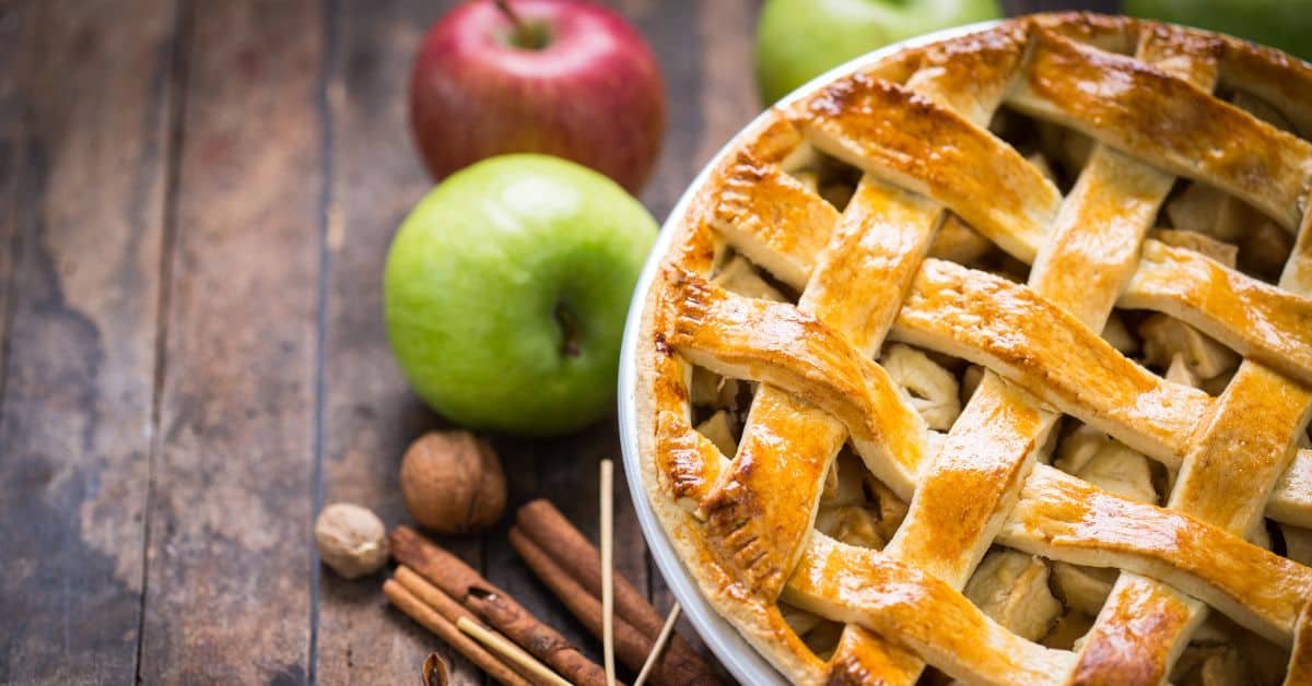 Delicious homemade apple pie with a golden lattice crust, surrounded by fresh red and green apples, cinnamon sticks, and whole spices on a rustic wooden surface.