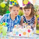 Two children in colorful party hats blow out candles on a festive birthday cake decorated with polka dots, surrounded by vibrant decorations and drinks at an outdoor celebration.