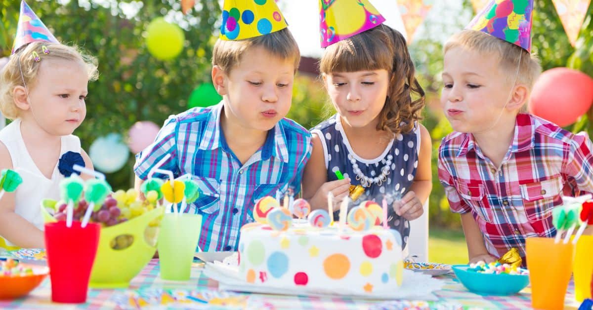 Two children in colorful party hats blow out candles on a festive birthday cake decorated with polka dots, surrounded by vibrant decorations and drinks at an outdoor celebration.