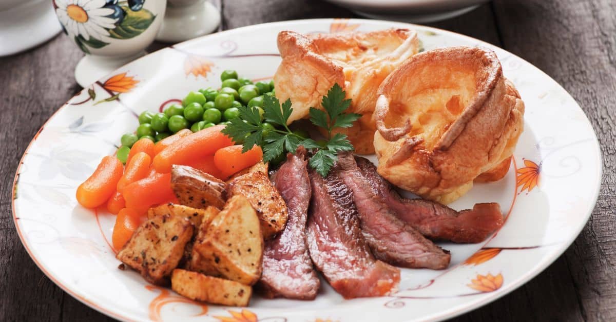 A delicious plate featuring tender slices of beef, roasted potatoes, vibrant green peas, and carrots, accompanied by a golden Yorkshire pudding and garnished with fresh parsley. Perfect for a hearty meal.