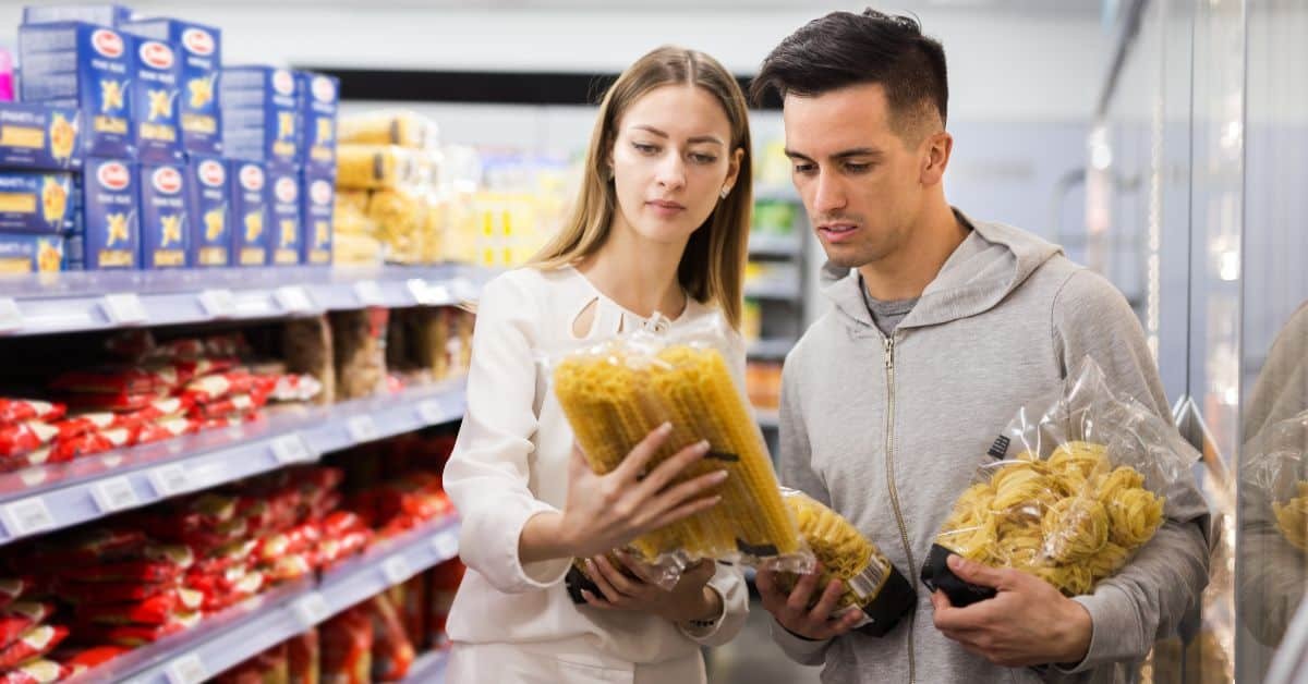 A couple shopping for pasta in a grocery store, examining different brands of pasta on the shelves.