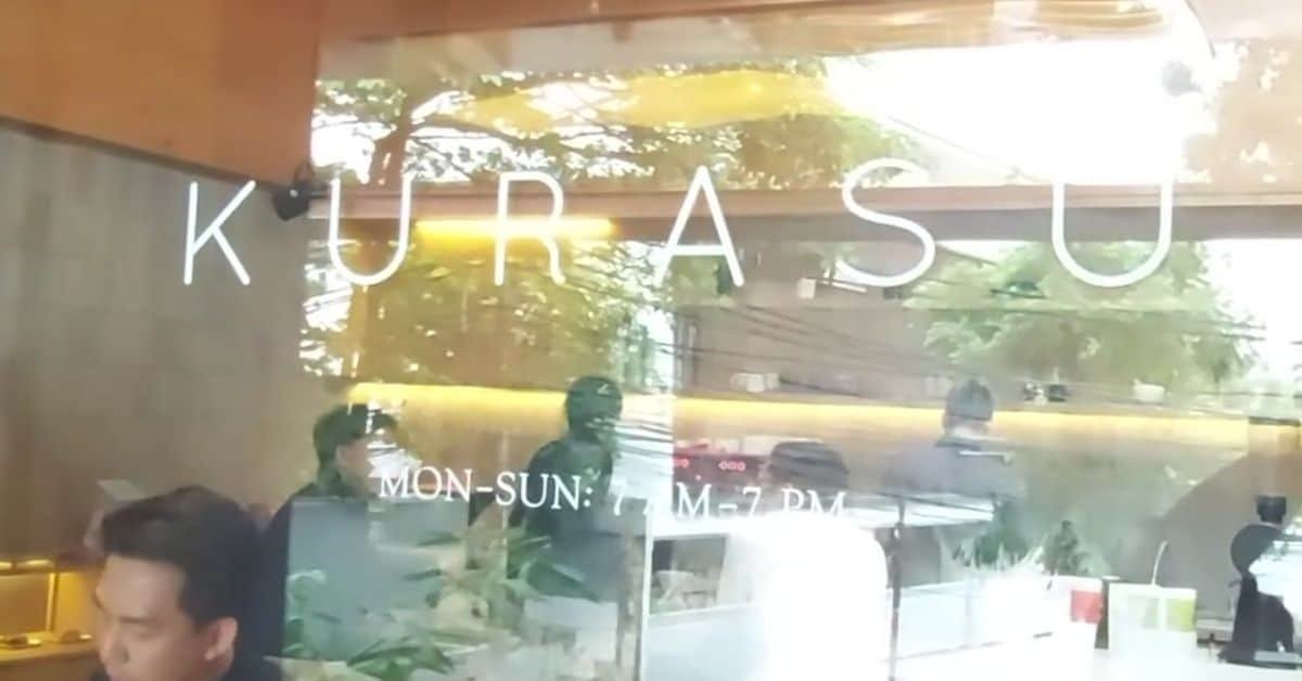 Glass storefront of a restaurant named "URAS," displaying operating hours from Monday to Sunday, with reflections of greenery in the background.