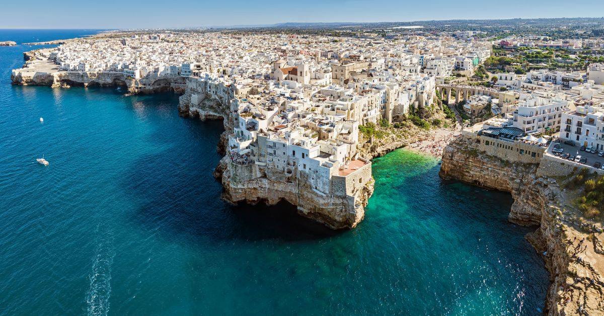 Aerial view of the coastal town of Polignano a Mare in Italy, showcasing whitewashed buildings perched on cliffs above turquoise waters, highlighting its stunning natural beauty and historic architecture.