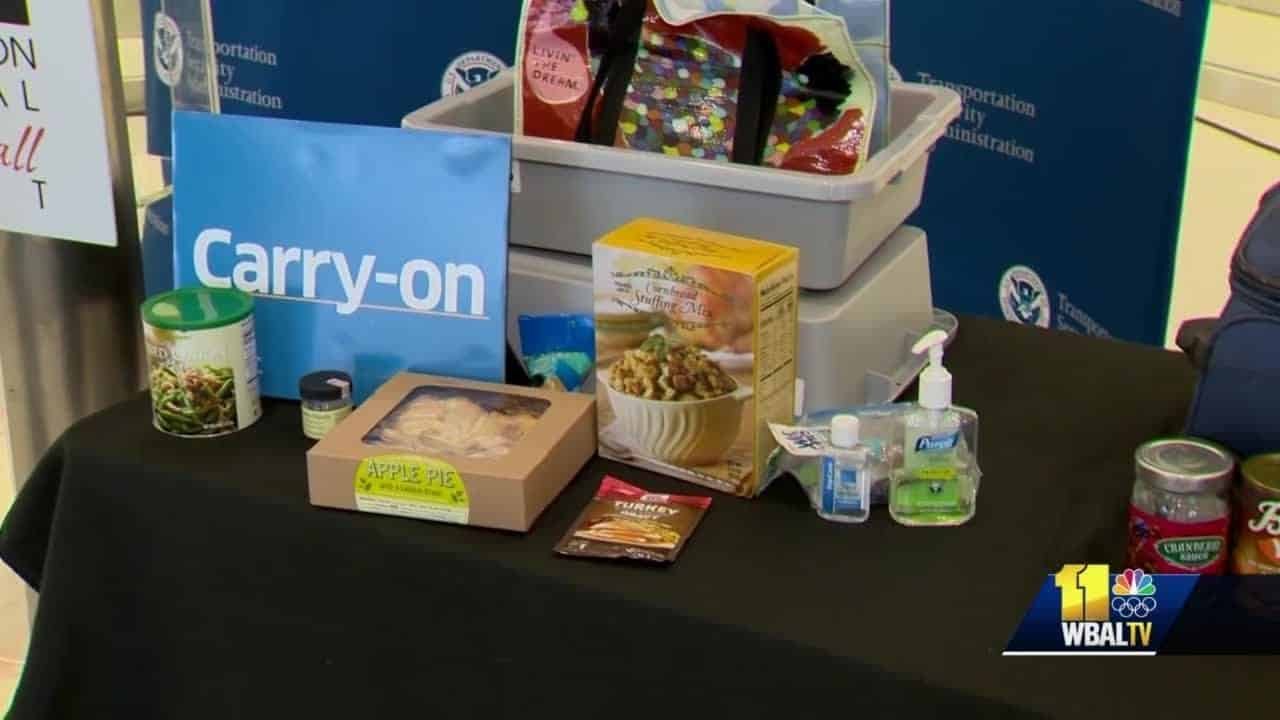 Display of travel essentials for carry-on luggage, including snacks, hand sanitizer, and toiletries, arranged on a table with a blue carry-on sign.