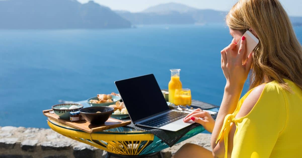 Person working on a laptop outdoors with a scenic ocean view, enjoying a breakfast spread and fresh juice, ideal for remote work and travel inspiration.