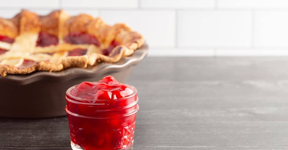 Homemade cherry pie filling in a glass jar, with a freshly baked cherry pie in the background on a gray countertop.