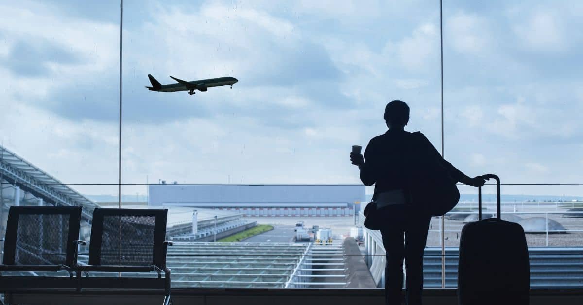 Silhouette of a person holding a coffee cup, looking out of an airport window as an airplane takes off against a cloudy sky.