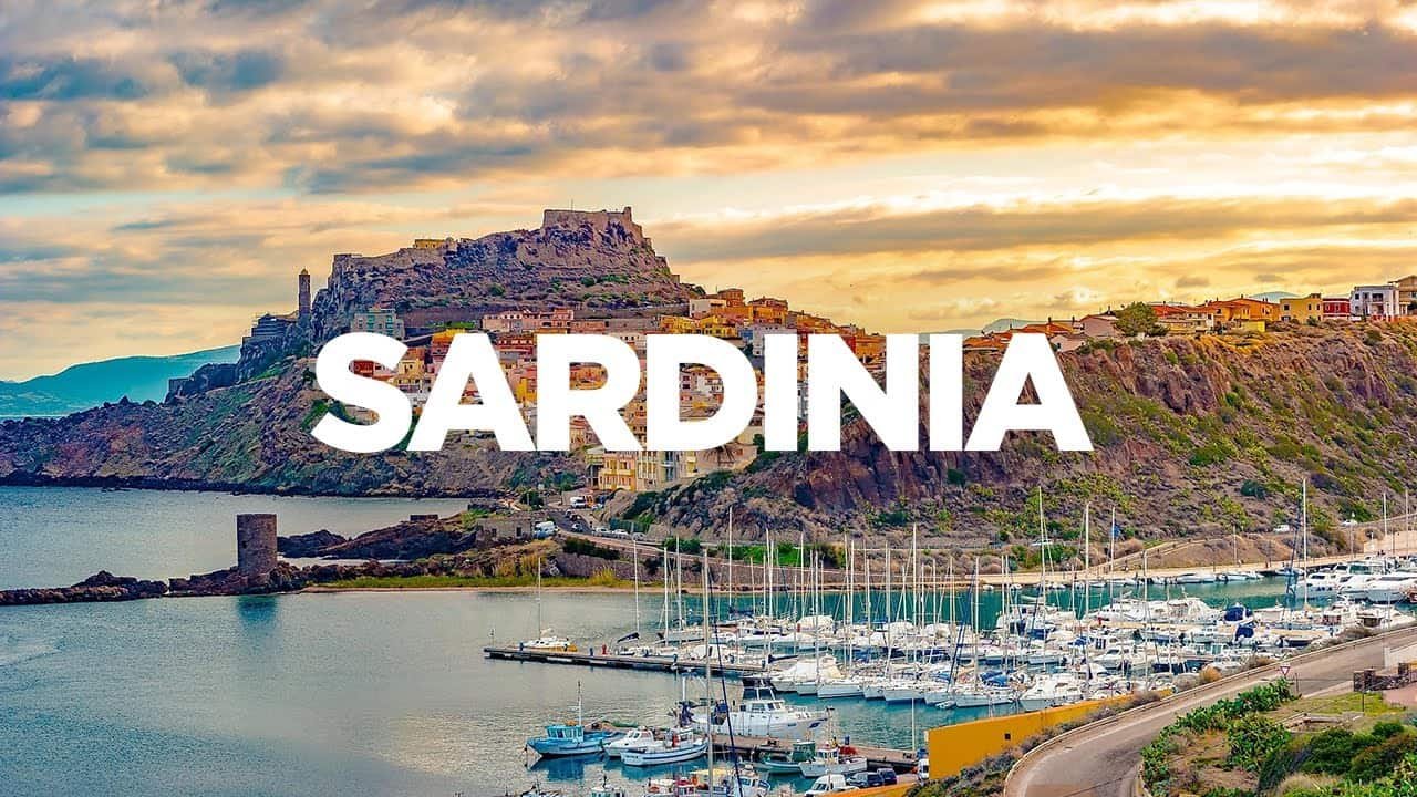 Scenic view of Sardinia featuring a marina with yachts and a mountainous coastline under a colorful sunset sky.