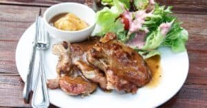 Grilled pork chop served with a side of mixed greens and a small bowl of mustard sauce, showcasing a delicious and hearty meal.