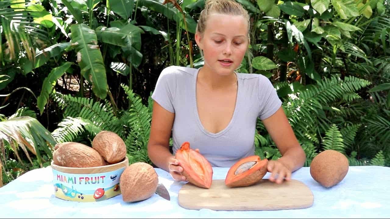 Woman demonstrating how to cut open cacao pods in a tropical setting, surrounded by fresh cacao fruits and a bowl.