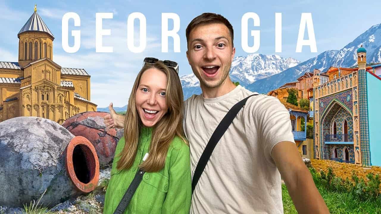 Young couple smiling for a selfie in Georgia, with scenic mountains and colorful buildings in the background.