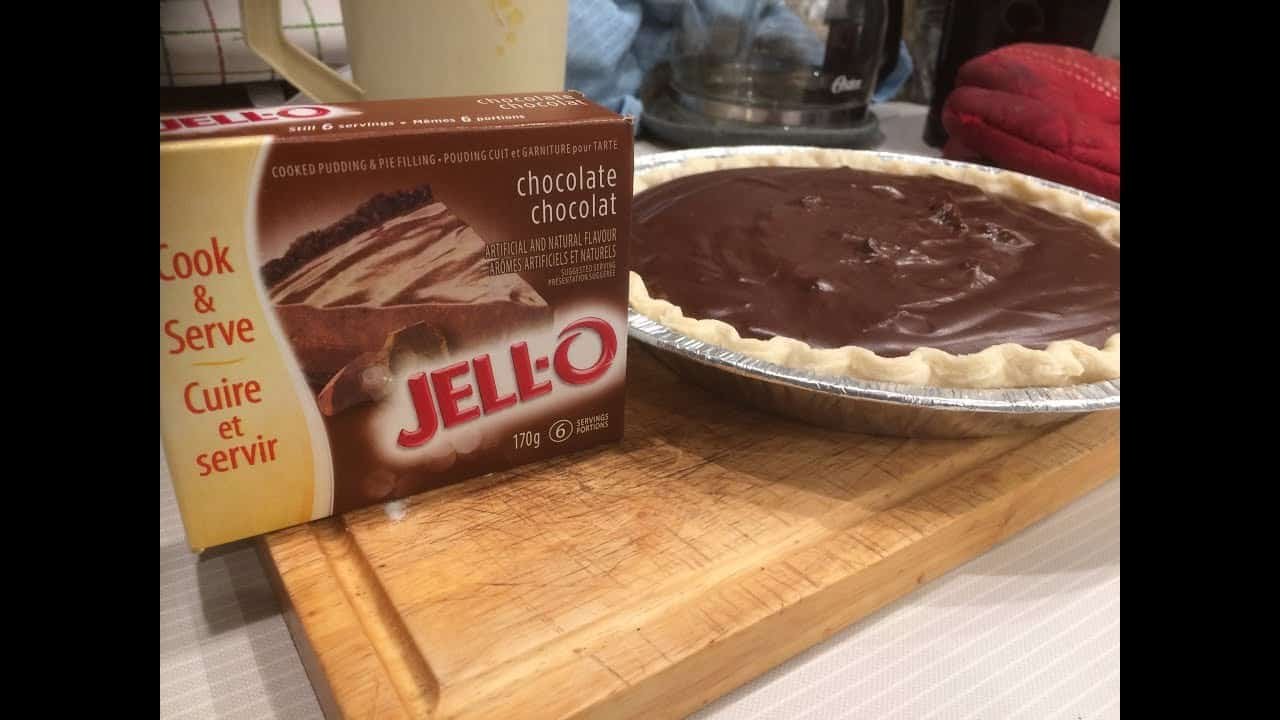 Box of JELL-O chocolate pudding mix next to a chocolate pudding pie on a wooden cutting board. The pie features a smooth chocolate filling in a baked crust, showcasing a homemade dessert.