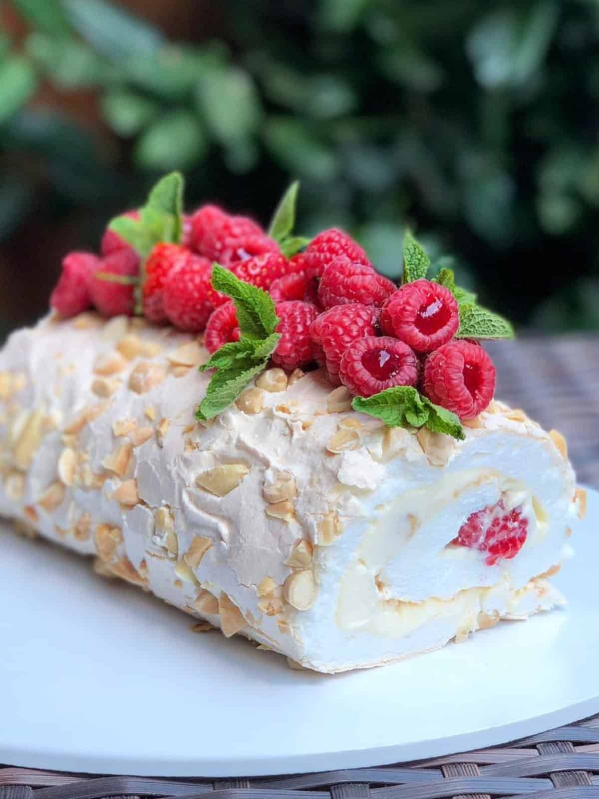 A beautifully decorated raspberry and almond meringue cake roll topped with fresh raspberries and mint leaves, set against a blurred green background. Perfect for dessert lovers and special occasions.