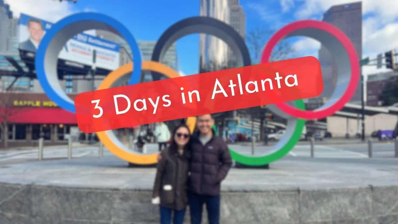 Couple posing in front of the Olympic rings in Atlanta, showcasing a travel guide theme titled "3 Days in Atlanta" against a clear blue sky.