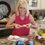 Cheerful woman in a kitchen, holding a slice of dessert, surrounded by various dishes and colorful ingredients on the counter, showcasing a vibrant cooking atmosphere.