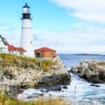 Portland Head Light lighthouse standing on rocky coast with ocean waves, surrounded by green grass and a clear blue sky.