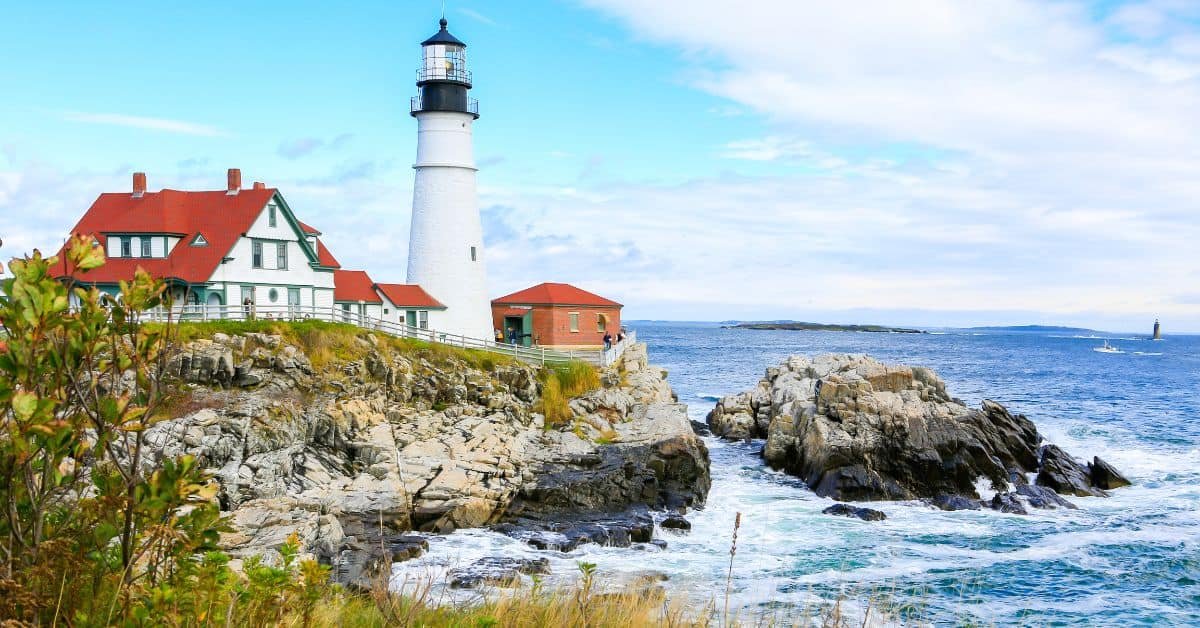 Portland Head Light lighthouse standing on rocky coast with ocean waves, surrounded by green grass and a clear blue sky.