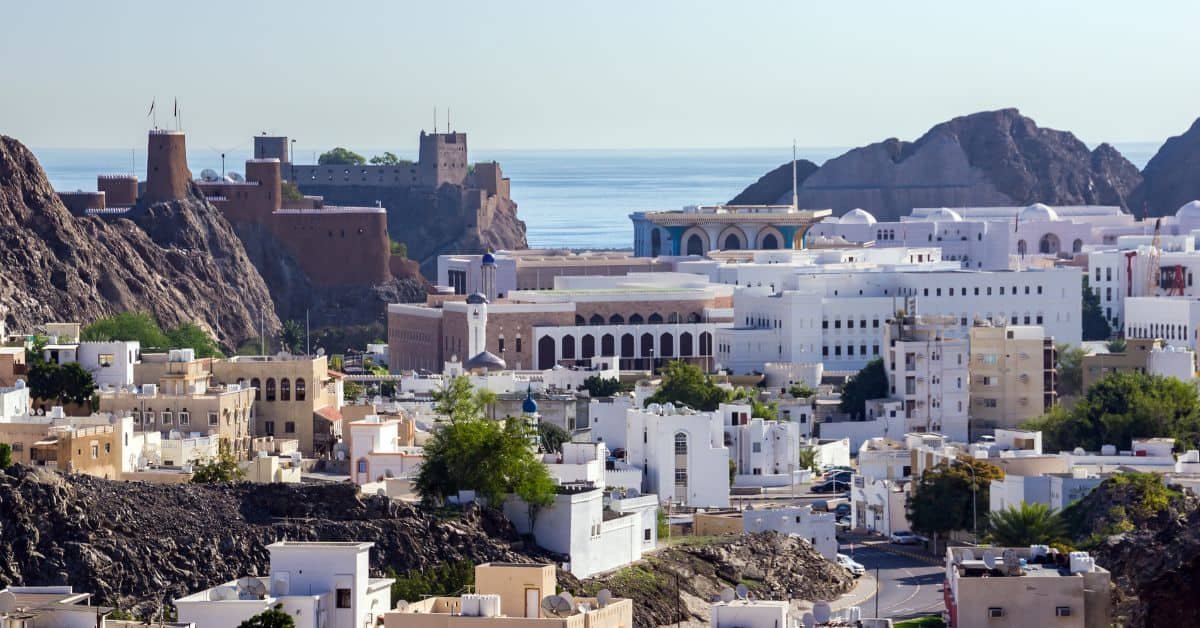 Panoramic view of a coastal city featuring traditional white buildings, a historical fort in the background, and a serene sea. The image captures the blend of modern architecture and historical landmarks, highlighting the scenic beauty of the area.