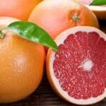 Fresh pink grapefruit halves and whole fruit with green leaves on a wooden surface, showcasing their juicy texture and vibrant color.