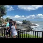 Family posing together at a scenic coastal viewpoint with lush greenery and ocean waves in the background under a bright blue sky.