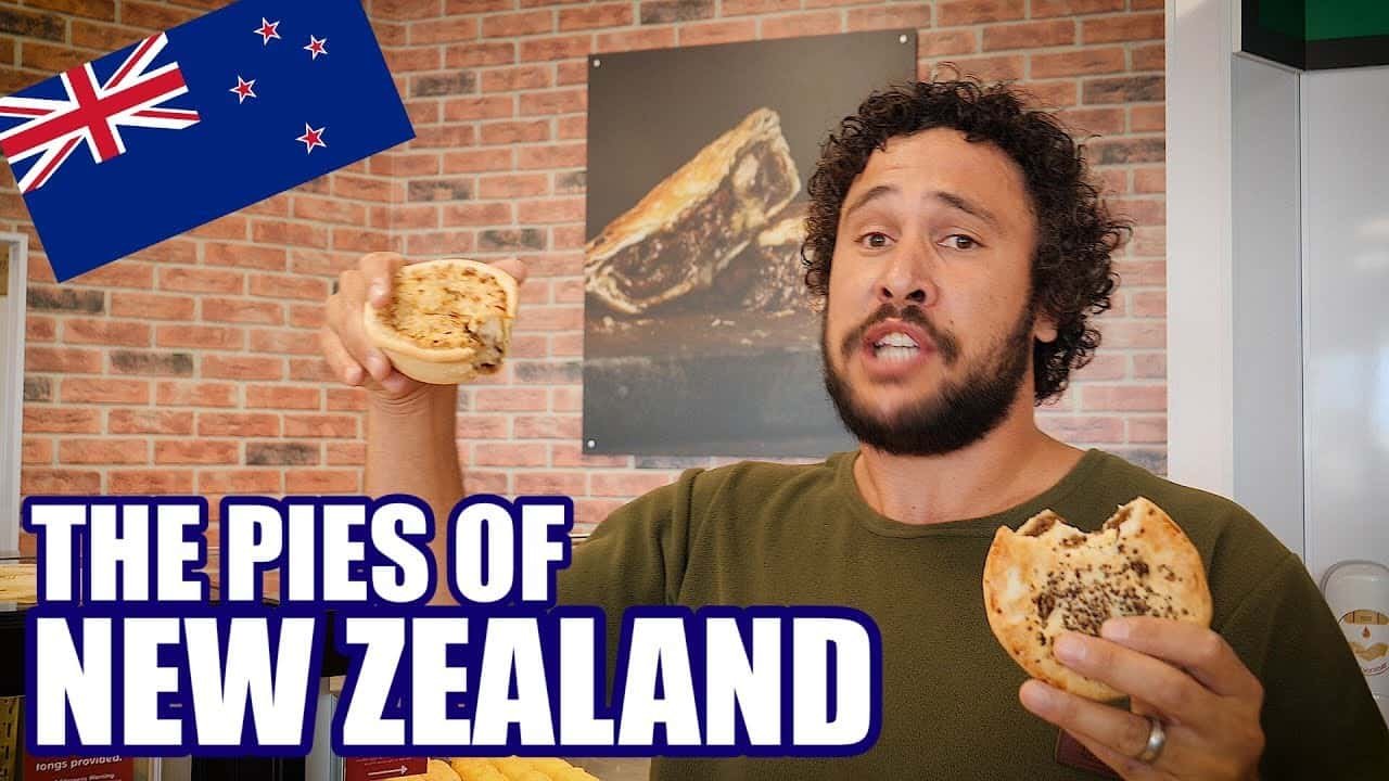 A man holding two unique food items from New Zealand, showcasing the country's culinary culture, with a backdrop featuring a Kiwi flag and a brick wall.