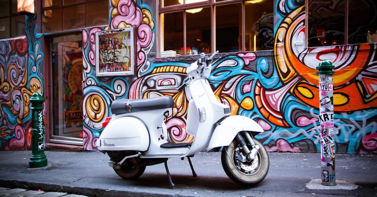 White vintage scooter parked in front of a colorful mural, showcasing vibrant street art and an urban setting.