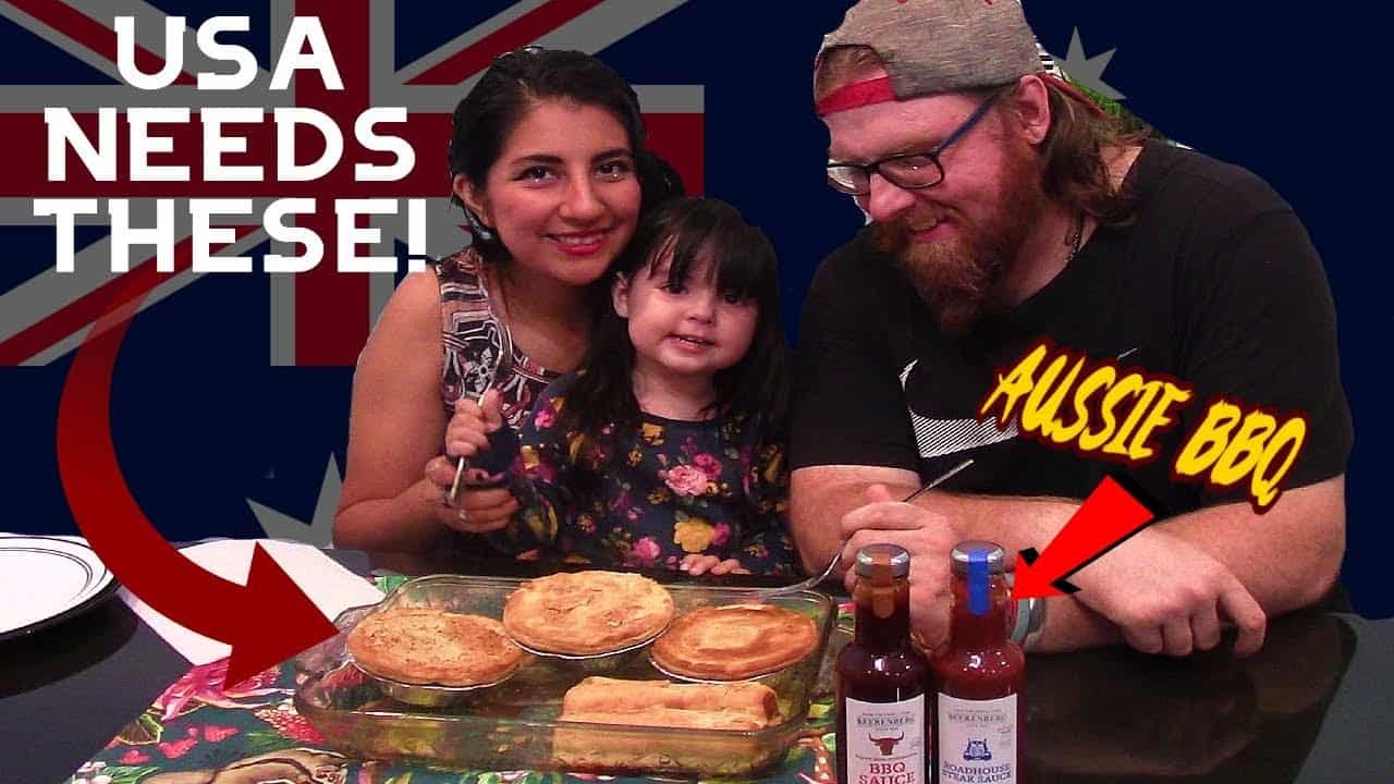 A family of three, including a woman, a young girl, and a man, smiles together in front of a table filled with various baked dishes and sauces, with a British flag in the background.