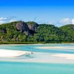 Scenic view of a tropical beach with white sand, crystal clear turquoise water, and lush green hills in the background under a bright blue sky. Ideal destination for relaxation and outdoor activities.