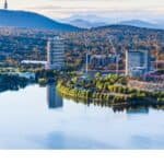 Australian National University vs University of Sydney: Which Should You Choose? Aerial view of a serene lake surrounded by modern buildings and lush greenery, with mountains in the background, showcasing a picturesque urban landscape.